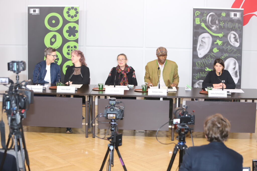 5 Personen sitzen hinter den Tischen des Podiums, drei Kameras sind auf sie gerichtet, i Hintergrund Dekorationen von ORANGE 94.o und Presseclub Concordia